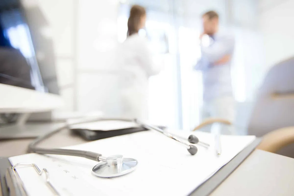 A stethoscope resting on a clinic desk while two clinicians talk in the background